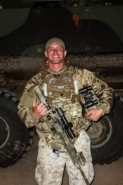 A muscular man in full tactical military uniform and camouflage gear poses with a rifle in front of a large vehicle at night.