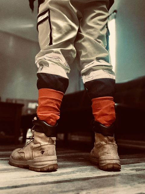 A low-angle view from behind showing a person wearing light cargo pants, bright orange socks, and scuffed tan leather work boots.