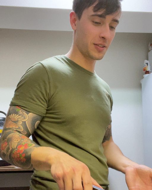 A muscular young man with a colorful sleeve tattoo wears a tight olive green t-shirt in a domestic setting.