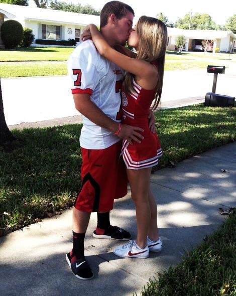 A couple in sports uniforms kisses on a sidewalk outside.