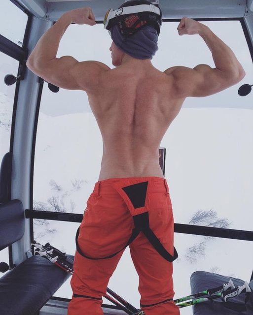 A shirtless muscular man in orange ski pants flexes his back and biceps while standing inside a gondola overlooking a snowy mountain.