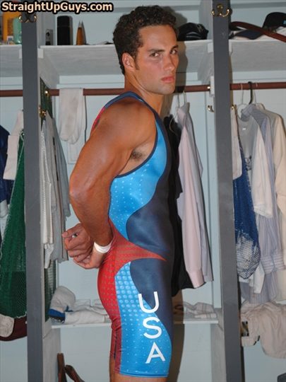 A muscular man in a tight red, white, and blue USA wrestling singlet stands in a locker room with his hands clasped behind his back.