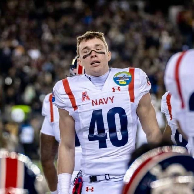 A muscular young man in a full Navy football uniform with eye black stands on a stadium field.