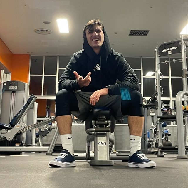 A young twunk in a hoodie, gym shorts, and compression leggings sits on a weight bench in a commercial gym.
