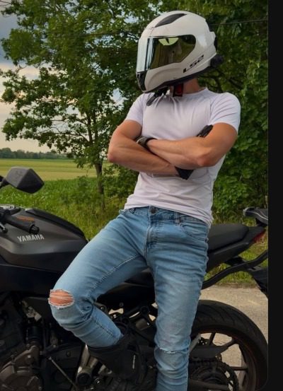 A man wearing a full-face motorcycle helmet and a tight white shirt leans against a Yamaha motorcycle in an outdoor setting.