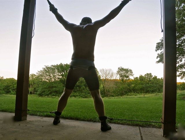 A muscular man wearing a wrestling singlet pulled down to his waist is restrained by his wrists and ankles in a spread eagle position on an outdoor porch.