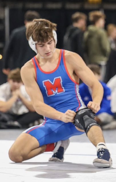 A lean, muscular young man in a blue and red wrestling singlet and headgear kneels on a mat to adjust his knee pad.