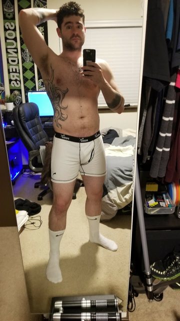A shirtless tattooed man poses in white boxer briefs and white crew socks for a mirror selfie in a bedroom.