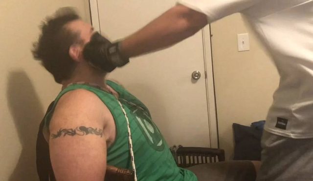 A man wearing a white t-shirt strikes a seated man in the face with a gloved hand indoors.