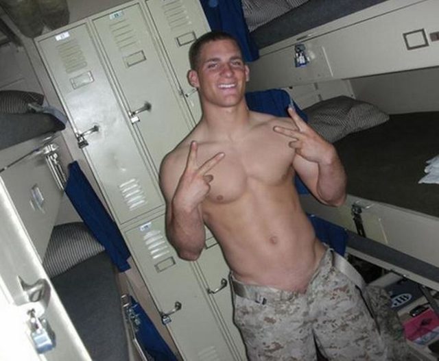 A muscular, shirtless young man in camo pants poses with peace signs in a military barracks setting with lockers and bunk beds.