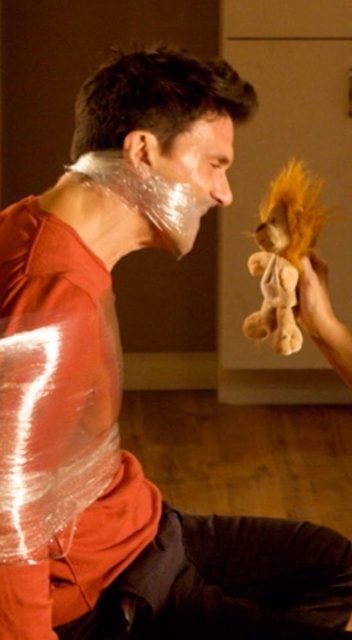 A man bound around his chest and lower face with clear plastic wrap looks at a small stuffed animal held in front of him.
