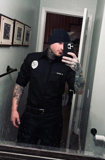 A heavily tattooed man in a black cop uniform and beanie takes a mirror selfie.