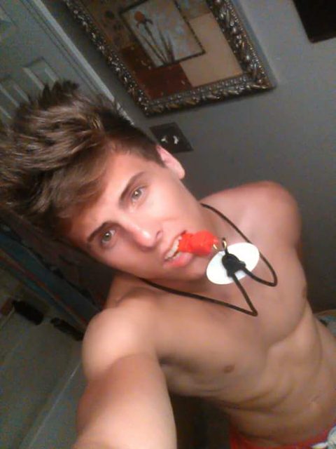 A smooth, shirtless twunk takes a high-angle amateur selfie while biting down on a red pacifier with a lanyard around his neck.