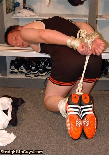 A man wearing a black wrestling singlet and athletic shoes is hogtied with rope on a locker room floor, resting his upper body against a bench.