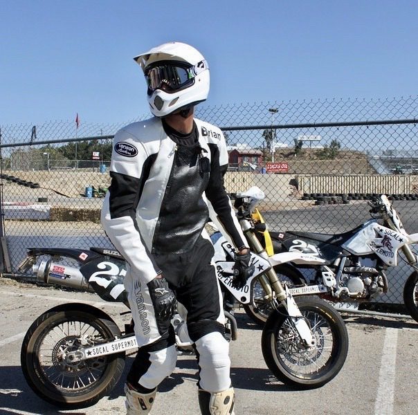 A man stands at a racing track wearing a full black and white leather motorcycle suit, helmet, and gloves.