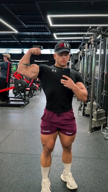 A muscular bodybuilder in a tight black t-shirt and maroon gym shorts takes a mirror selfie while flexing his bicep in a gym.