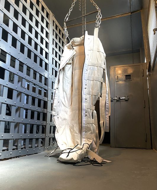 A person is fully encased and suspended in a heavy canvas restraint bag within a metal-barred jail cell or dungeon.