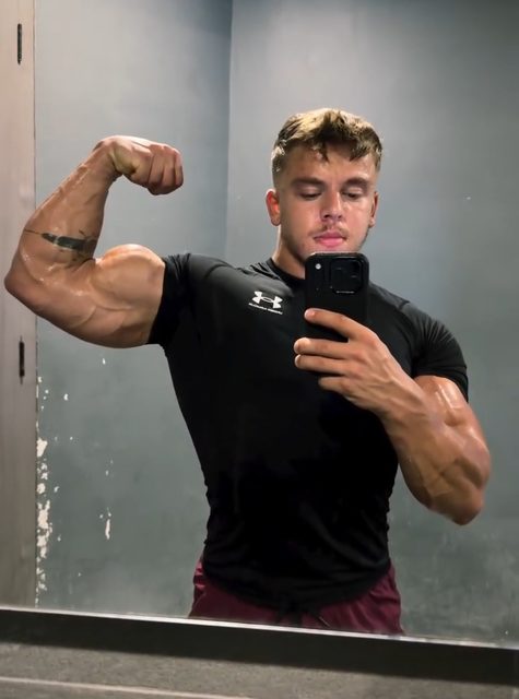 A muscular young man in a black compression shirt and gym shorts flexes his bicep for a mirror selfie in a locker room.