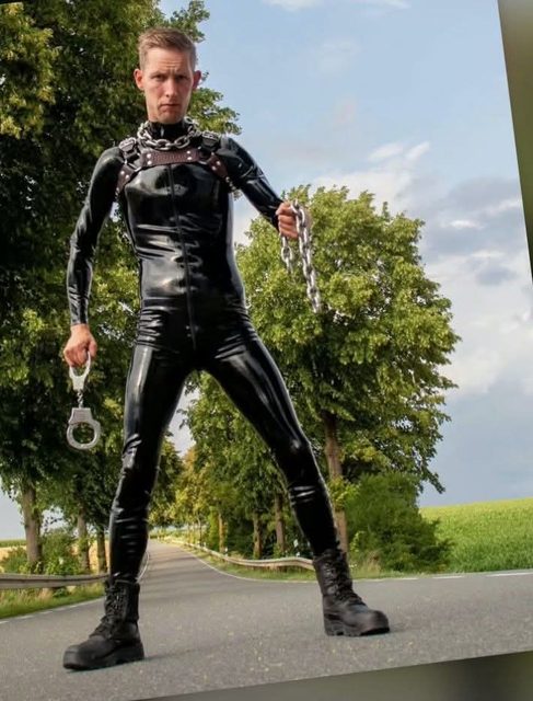 A man wearing a full black latex catsuit, a leather harness, and heavy metal chains stands on a public road holding handcuffs.