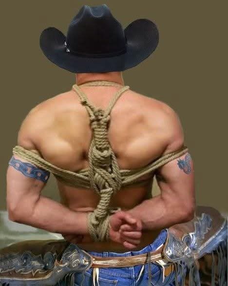A muscular man seen from behind wears a cowboy hat and jeans while his arms are bound behind his back with thick rope.