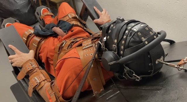 A man in an orange prison jumpsuit is immobilized by multiple leather straps and a studded leather hood equipped with headphones.