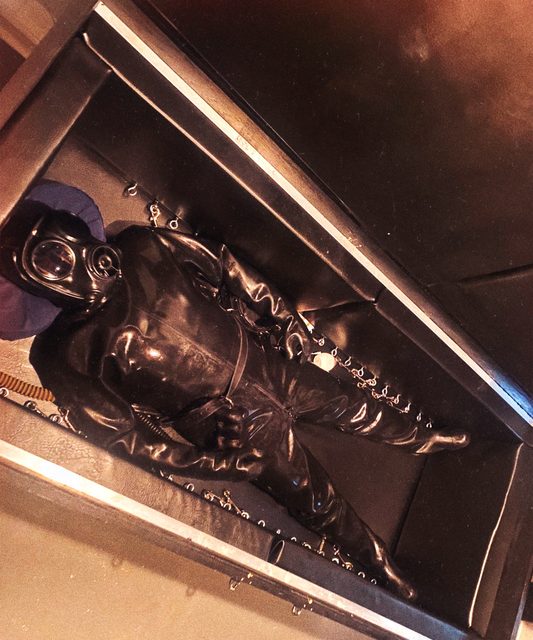 A person wearing a full black latex suit and gas mask lies inside a padded confinement box.