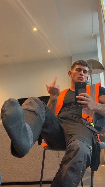 A muscular man in construction gear and a hi-vis vest sits in a chair, extending a socked foot toward the camera while taking a selfie.