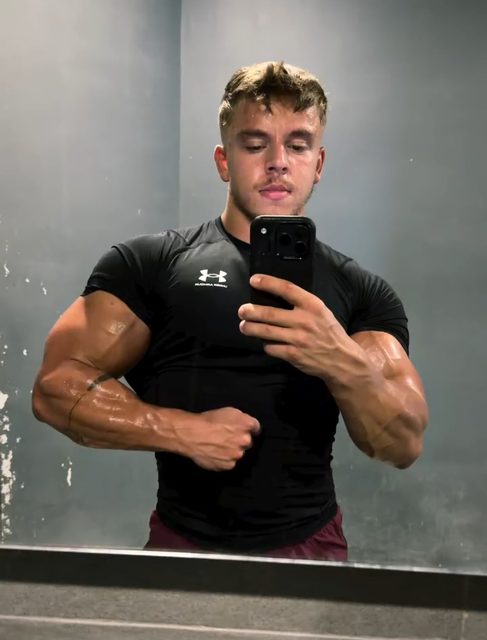 A muscular young man in a tight black compression shirt flexes his biceps for a mirror selfie in a gym locker room.