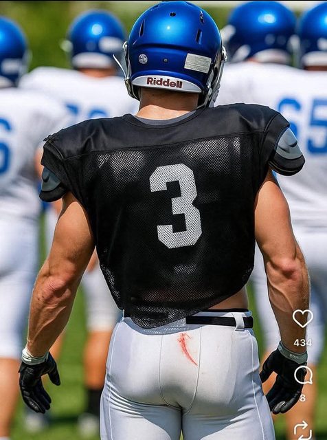 A muscular football player in full gear and tight white pants stands on an outdoor field.