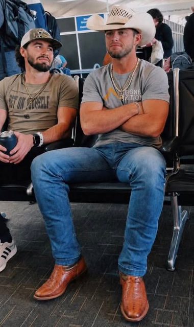 Two masculine men sit in a public waiting area, featuring one man in a cowboy hat and tight denim jeans sitting with his legs spread.