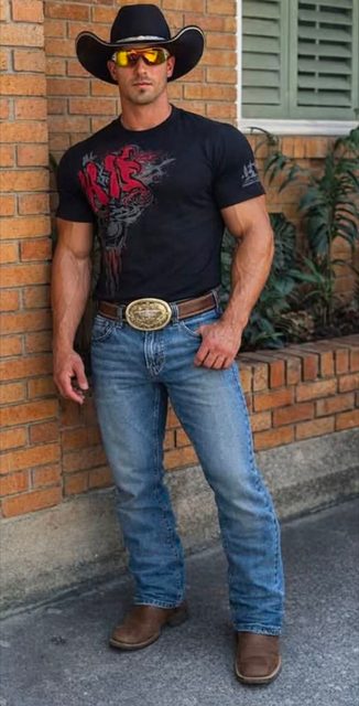 A muscular man with vascular arms poses outdoors wearing a black cowboy hat, graphic t-shirt, denim jeans, and a large trophy belt buckle.
