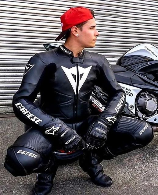 A smooth-faced man in full black Dainese motorcycle leathers and a red cap squats in front of his bike and a metal shutter.