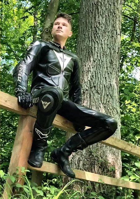A young man in a full Dainese leather motorcycle suit, gloves, and boots sits on a wooden fence in a wooded area.
