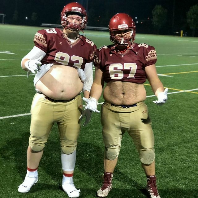 Two men in full American football gear pose on a grass field at night with their jerseys pulled up to expose their bellies.