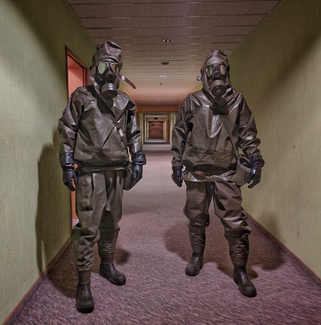 Two individuals wearing full olive-drab hazmat suits and gas masks stand in a long hotel hallway.