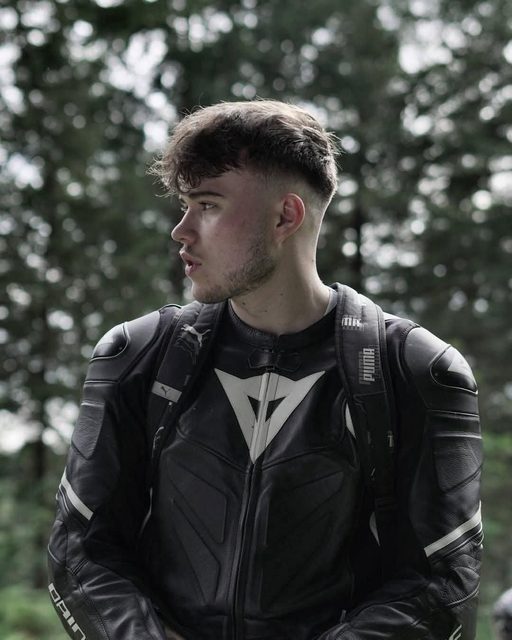A young man with a fade haircut and light beard wearing a black and white leather motorcycle jacket and a backpack outdoors.