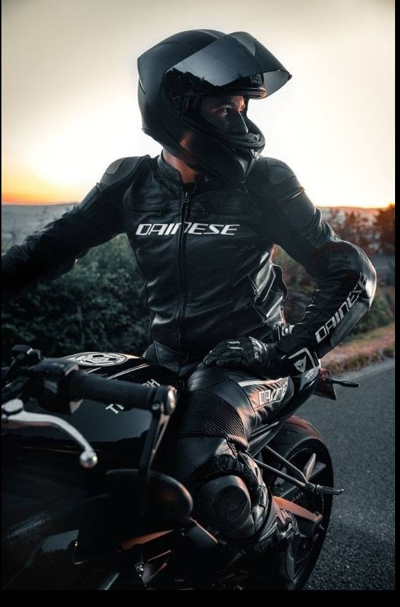 A rider dressed in a full black leather motorcycle suit and helmet poses on a sports bike outdoors at sunset.