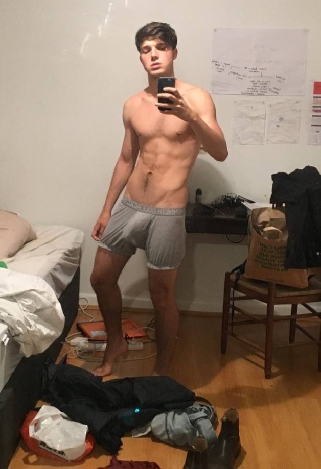 A shirtless twunk with defined abs poses for a bedroom mirror selfie wearing grey boxer briefs with a visible bulge.