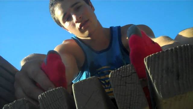 A young man in a blue tank top presents his feet in red socks to the camera outdoors.