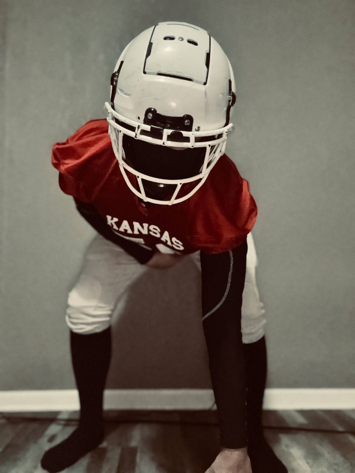 A man posing in a full Kansas football kit including a white helmet, red jersey, and compression sleeves.