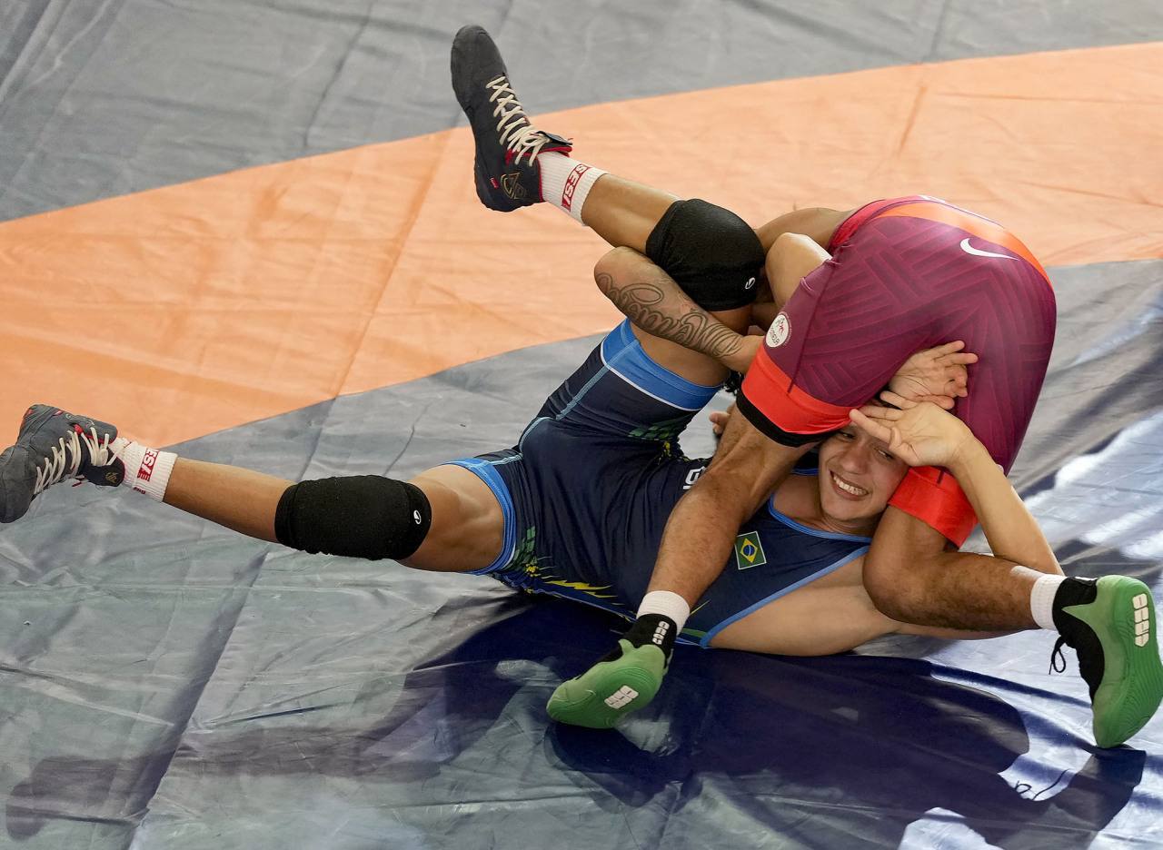 Two young athletes in spandex wrestling singlets grapple on a mat, with one wrestler pinning the other in a leg hold.