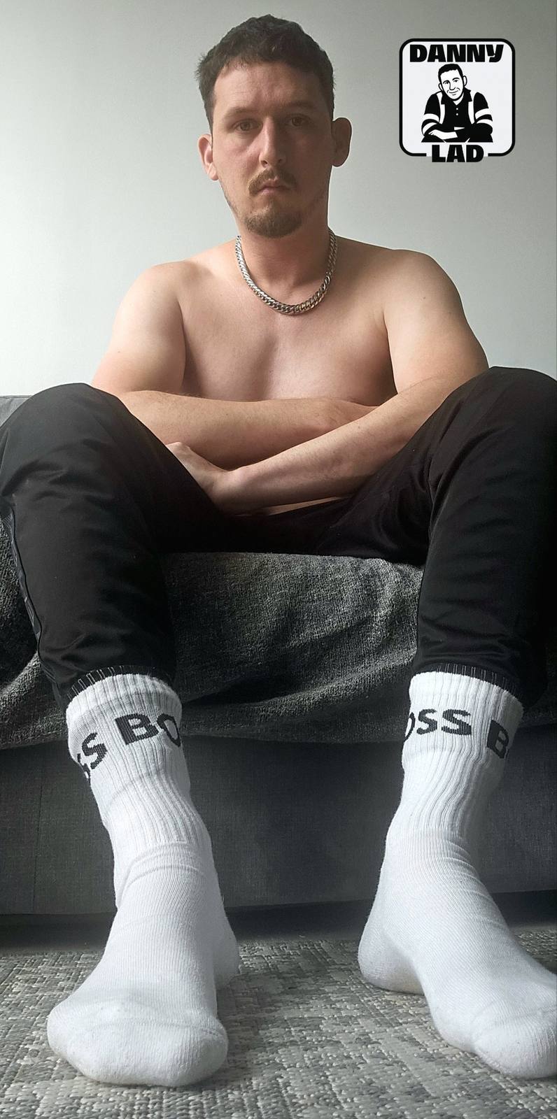 A shirtless man with a goatee and silver chain sits on a couch wearing black track pants and white BOSS crew socks.
