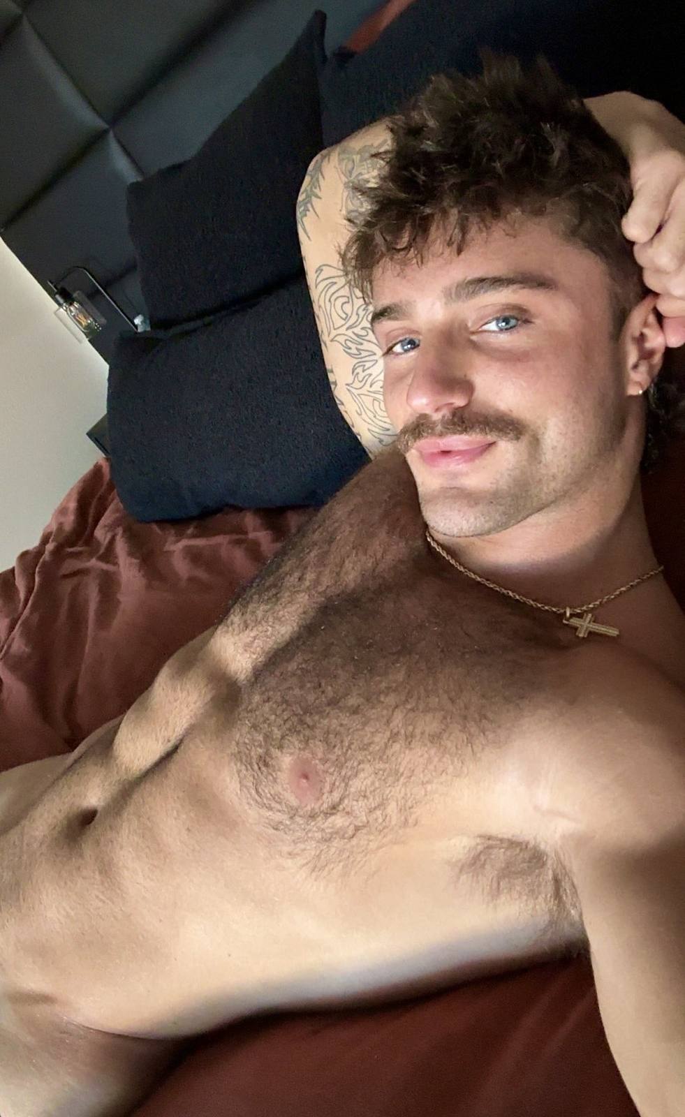 A shirtless, hairy-chested man with a mustache and arm tattoo lies in bed looking directly into the camera.