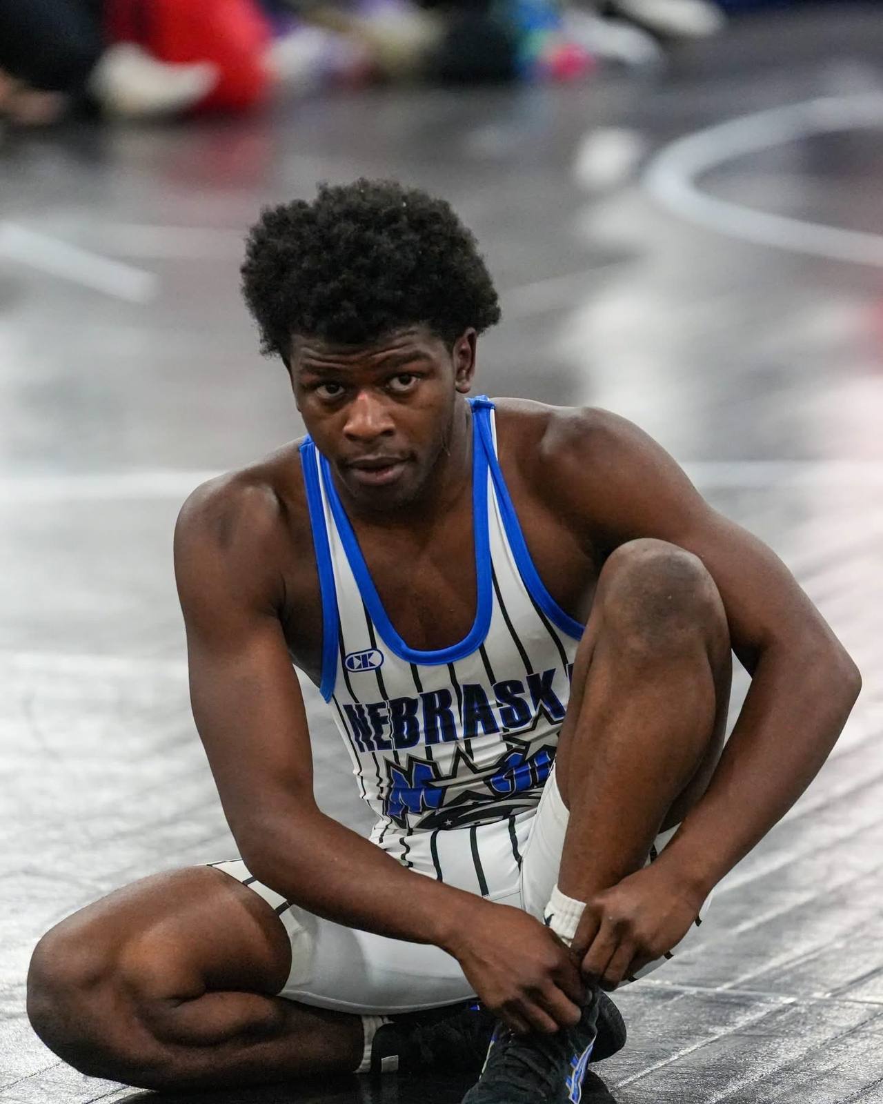 A muscular young man in a Nebraska wrestling singlet sits on a mat while adjusting his gear.