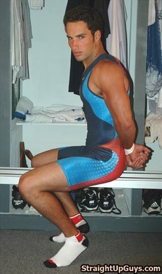 A muscular jock sits in a locker room wearing a blue and red wrestling singlet and low-cut socks.