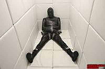 A person fully enclosed in a black latex gimp suit sits restrained on the floor of a white padded cell.