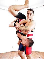 Two muscular men in wrestling singlets engage in a grappling session where one man lifts the other upside down.