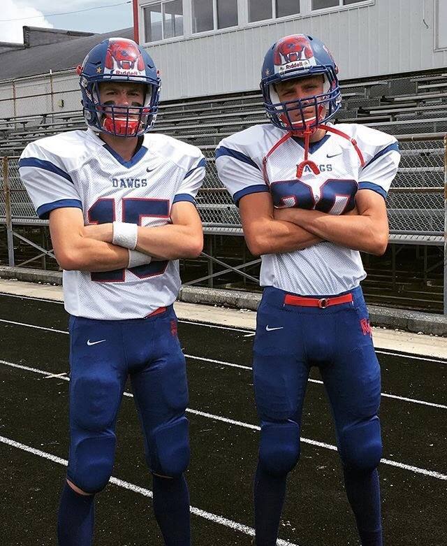Two athletic young men in full football gear and helmets pose with arms crossed on an outdoor track.