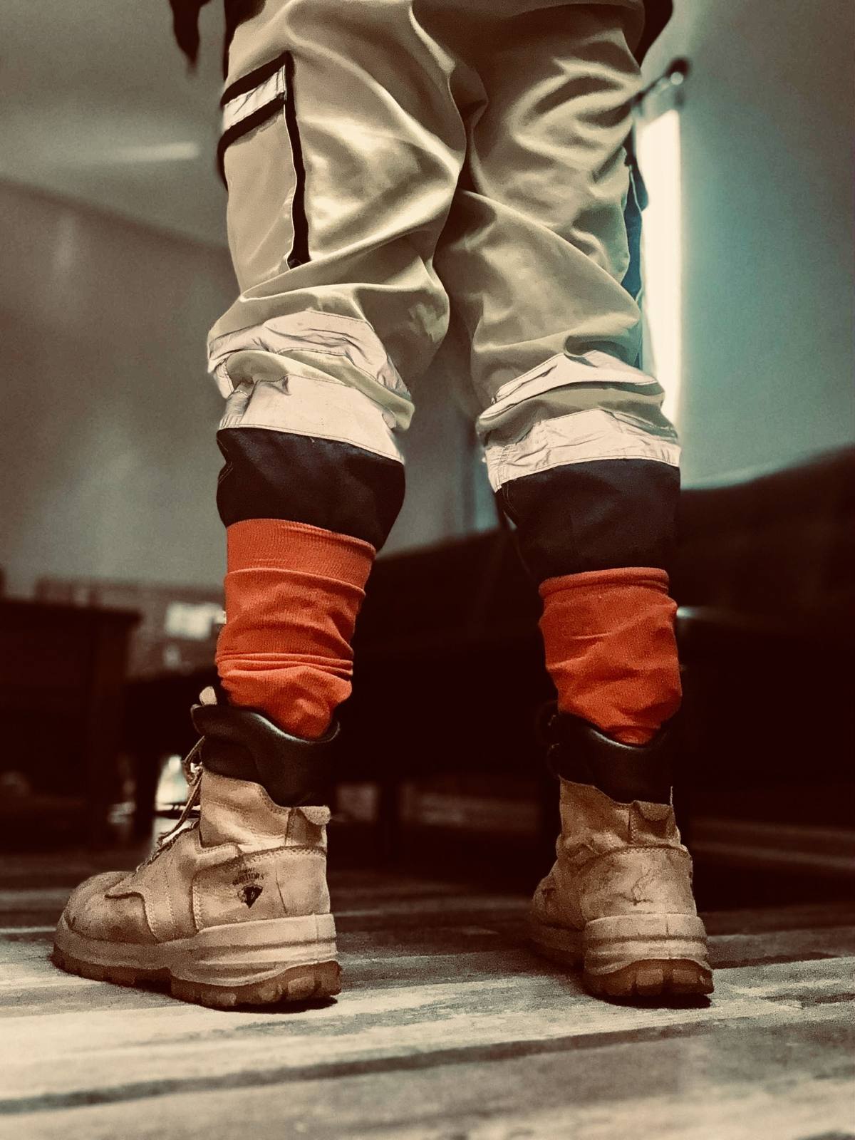 A low-angle view from behind showing a person wearing light cargo pants, bright orange socks, and scuffed tan leather work boots.
