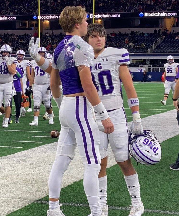 Two young athletic football players in tight purple and white uniforms converse on the field after a game.